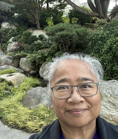 Headshot of grey haired woman wearing glasses, smiling at camera, with large rocks and bushes in the background.