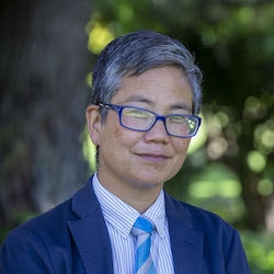 Amy Sueyoshi standing outdoors in front of a tree wearing a coat and tie, headshot
