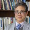 Amy Sueyoshi in front of a bookshelf in coat and tie, headshot