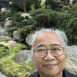 Headshot of grey haired woman wearing glasses, smiling at camera, with large rocks and bushes in the background.