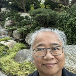 Headshot of grey haired woman wearing glasses, smiling at camera, with large rocks and bushes in the background.
